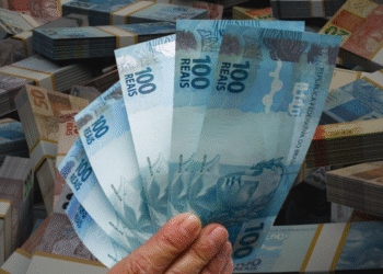 Mão segurando várias cédulas de R$ 100 com pilhas de dinheiro ao fundo.