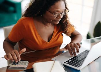 Mulher madura usando notebook e calculadora para organizar as finanças em casa, com caderno e caneta sobre a mesa