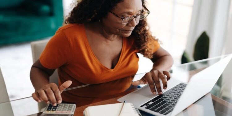 Mulher madura usando notebook e calculadora para organizar as finanças em casa, com caderno e caneta sobre a mesa