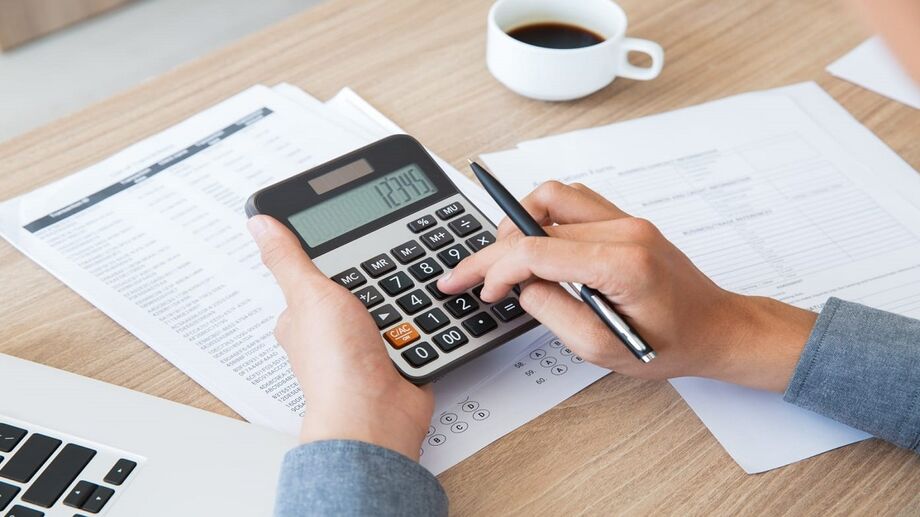Pessoa usando calculadora e segurando caneta para organizar finanças pessoais com documentos e laptop ao fundo
