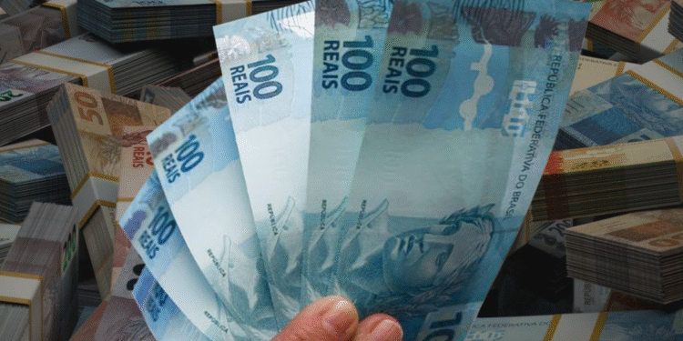 Mão segurando várias cédulas de 100 reais com pilhas de dinheiro ao fundo.