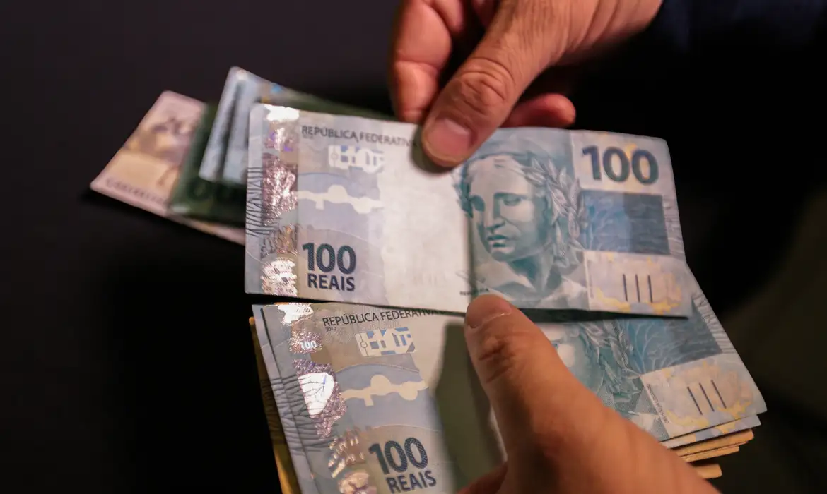 Mãos contando várias cédulas de 100 reais em ambiente escuro.