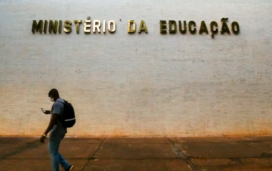 Fachada do Ministério da Educação com uma pessoa caminhando, usando máscara e olhando para o celular.
