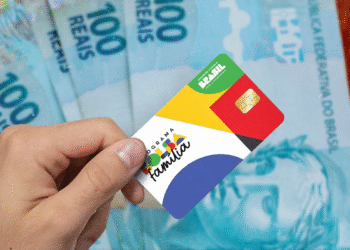 Cartão do Bolsa Família sendo segurado por uma mão com notas de 100 reais ao fundo