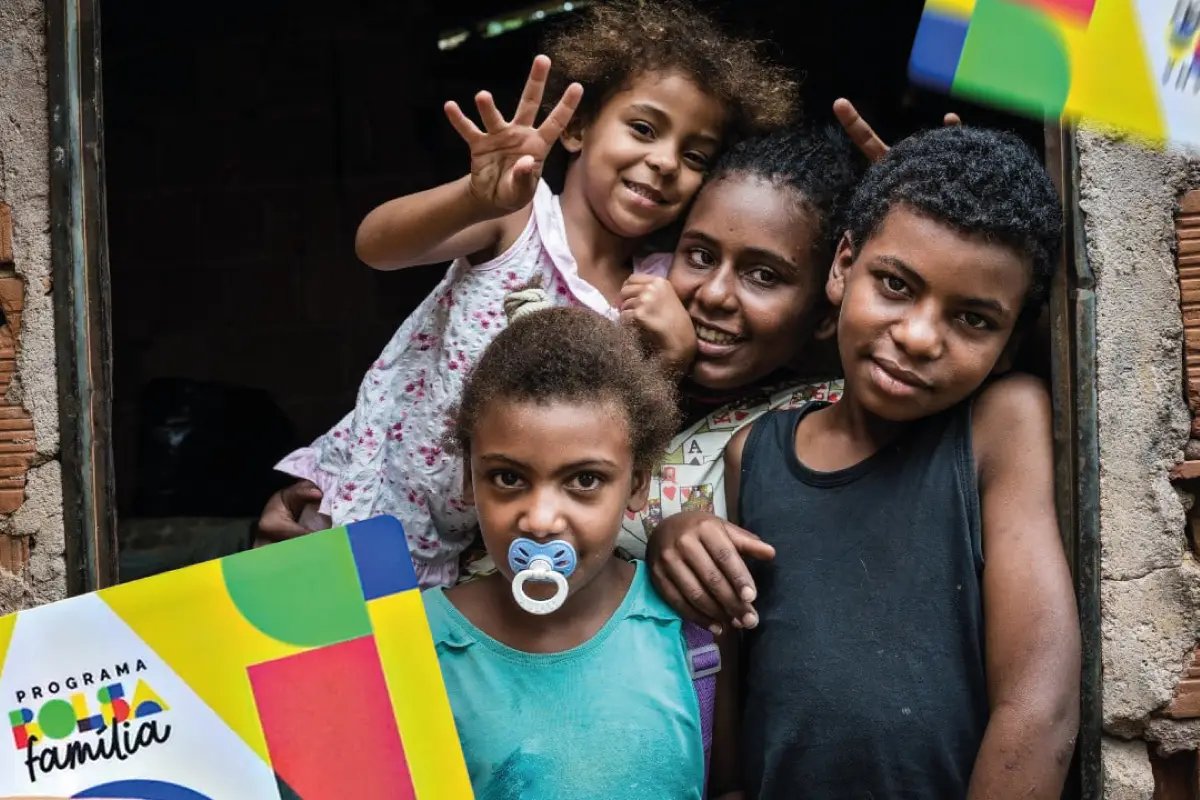 Crianças sorrindo, com destaque para um cartão do programa Bolsa Família, simbolizando novos grupos prioritários.