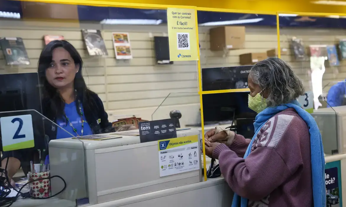 Funcionária de agência dos Correios assistindo cliente no balcão durante o processo de adesão a serviços.