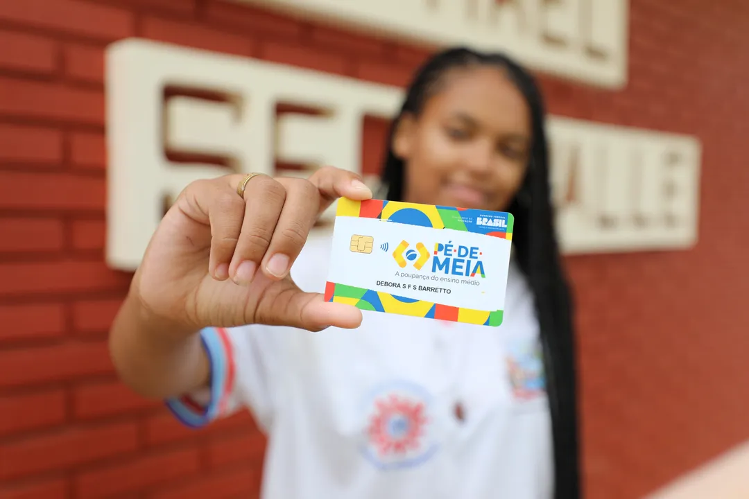 Cartão do programa Pé-de-Meia em agosto sendo segurado com notas de 100 reais ao fundo, representando o pagamento do incentivo financeiro aos estudantes.