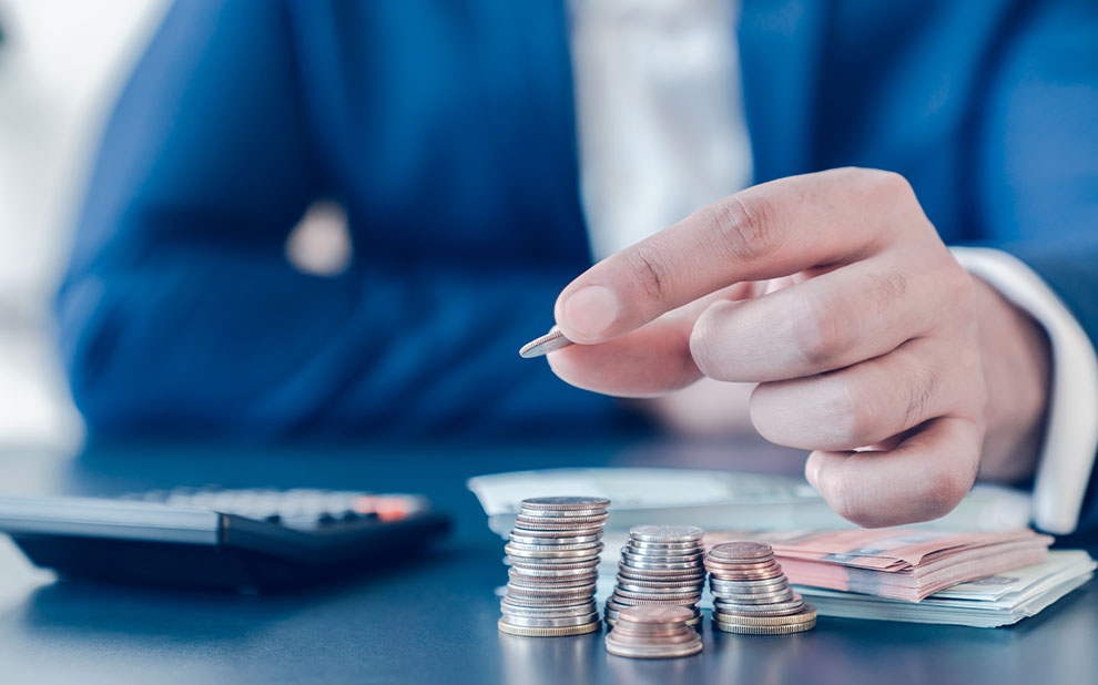 Homem de terno ajustando moedas sobre pilha de dinheiro com uma calculadora e cédulas de fundo, ilustrando um empréstimo. 
