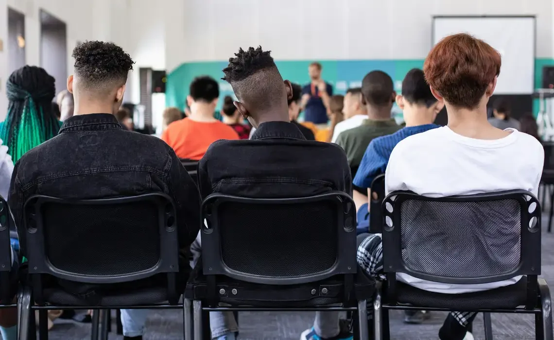 Três estudantes sentados, de costas, assistem a uma palestra, destacando a importância da educação e do incentivo a matricula.