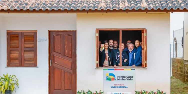 Grupo de pessoas sorrindo na janela de uma nova casa do programa Minha Casa, Minha Vida, mostrando a chave da moradia. Moradias pelo governo.